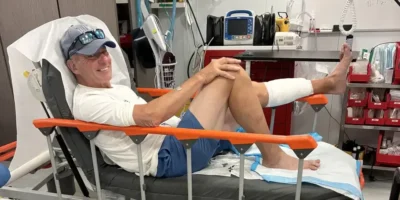 John Schweizer assis sur un lit d'hôpital après la morsure de requin aux Bahamas, la jambe gauche bandée