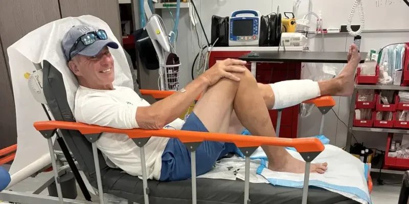 John Schweizer assis sur un lit d'hôpital après la morsure de requin aux Bahamas, la jambe gauche bandée